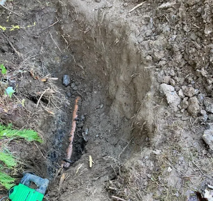 An orange drainage pipe lying in an open trench in the ground, surrounded by soil and roots, suggesting a drain repair.